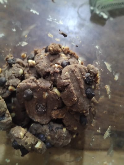 The image presents a close-up view of a pile of small, dark brown cookies or raw cookie dough pieces, densely packed within a clear container. The cookies are irregular in shape and size, displaying a coarse, crumbly texture. Embedded within the chocolate-flavored dough are numerous dark, small chocolate chips and lighter-colored chopped nuts, possibly peanuts or cashews, which are distinctly visible on the surface of many pieces. Some cookies exhibit faint indentations or ridges, suggesting they were shaped or pressed.

The container holding the cookies appears to be clear plastic or glass, with visible smudges, dust, or cookie crumbs adhering to its inner surface, particularly around the edges and in the blurred foreground. The background, especially the upper left quadrant, is out of focus but reveals a green object with some indistinct white text. This object appears to be part of another container or packaging. There is no activity taking place, as the image is a static shot of food. No specific time of day, weather, or emotions are discernible. The scene implies an indoor setting, likely a kitchen or pantry, in Gampongbaro, Indonesia.