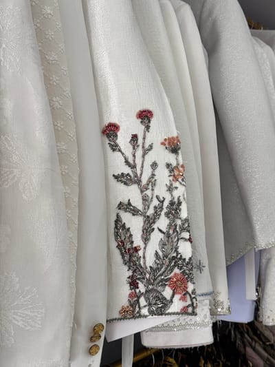 The image shows a close-up, eye-level shot of several white garments hanging in what appears to be a store or closet. The garments are made of different fabrics, with some featuring intricate embroidery and beadwork. One garment prominently displays a floral embroidery design in shades of red, pink, silver, and grey along the sleeve. Another garment on the left has a subtle, embossed floral pattern. Visible on the lower part of one garment are three gold-toned buttons. The background consists of more hanging garments, with some appearing to be light blue and other shades of white. The overall scene is indoors, with the lighting suggesting a well-lit environment, possibly artificial. There are no people visible in the image. The location in Tanjungmeru, Indonesia, is not discernible from the visual information provided.