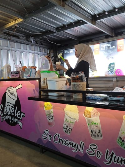 An indoor or semi-open food stall, likely in Sekopek Wetan, Indonesia, is depicted, specializing in a creamy dessert called "Es Teler." Two women are working behind a counter with a black top and a vibrant pink and purple branded front. The woman on the right, wearing a light-colored hijab and a black long-sleeved shirt, is actively preparing a drink or dessert, scooping ingredients from a large white container into a clear plastic cup. Another woman, also in a hijab, is partially visible behind other containers on the left side of the counter.
The stall's branding "Es Teler creamy yummy" is prominent on the counter, along with the slogan "So Creamy!! So Yummy" (partially visible). Illustrated cups filled with colorful desserts are also part of the counter design. Above the counter, menu boards display prices and options: "ORIGINAL 12 K" (Rupiah), "TOPING ICE CREAM LARGE 15 K", and topping choices like "MUTIARA - CREAMER - ALPUKAT", "SELASIM - ICE CREAM". A small sign on the left reads "ORDER & PAY HERE" and "Pesan & Bayar Di sini". A white fan is placed on the left side of the counter, and a security camera is mounted on the corrugated metal ceiling structure, which also has exposed wiring. A container on the counter has the text "STAN 1221". The overall setting suggests a casual food preparation area, possibly within a market or a small eatery.