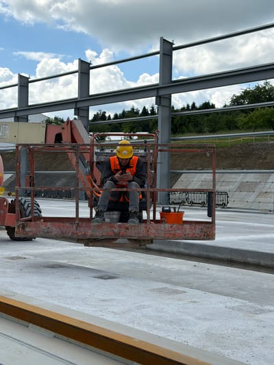 In Wilkowisko, Poland, a construction worker wearing a yellow hard hat and an orange safety vest sits in the basket of a cherry picker. The worker is looking down at a mobile phone in their hands. The cherry picker is positioned on a concrete surface, with a large steel structure under construction in the background. The sky is partly cloudy, with white clouds against a blue backdrop. Trees and a hillside are visible behind the steel structure.