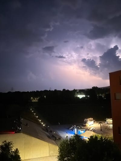 The image depicts a dramatic nighttime scene in Glattbrugg, Switzerland, characterized by a powerful lightning strike illuminating the sky. Dark, ominous storm clouds dominate the upper portion of the frame, with a brilliant flash of lightning emanating from the center-left, casting a pinkish hue across the cloud formations and the landscape below.

In the foreground, a modern, reddish-brown building juts into the frame from the right. Below and to the left of the building, a large, sloped roof structure is visible. This structure is illuminated by a series of small, bright lights that create a pathway along its surface. Several individuals are gathered on this roof, appearing to observe the storm. Some are standing, while others are seated or leaning. A red light is visible on the far left of this roof area.

Beyond the roof structure, the ground slopes downwards and away. This area is sparsely populated with some low-lying trees and vegetation. A few food trucks and stalls are set up, with lights illuminating their interiors and immediate surroundings. One food truck, with its service window open, is prominently lit. Picnic tables and benches are scattered around this area, suggesting a public gathering or event. Further in the distance, a dark, dense line of trees forms the horizon, with scattered lights indicating buildings or streetlights within a town or city. Two tall power pylons are visible in the far distance on the left and right edges of the frame, silhouetted against the stormy sky. The overall impression is one of a fleeting, awe-inspiring natural event observed from an urban setting.