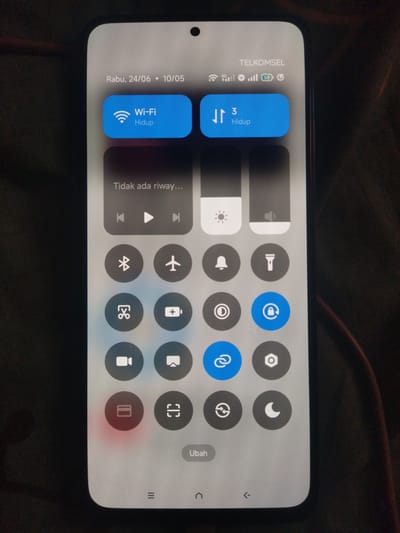 This is a media file showing a close-up of a smartphone screen displaying the control center. The screen is held in a portrait orientation against a dark, textured background. The time is indicated as "Rabu, 24/06 - 10/05," which translates to Wednesday, June 24th, 10:05 AM. The phone's carrier is displayed as "TELKOMSEL."

The control center offers several toggles and controls. Prominently displayed are two large blue buttons: one for "Wi-Fi Hidup" (Wi-Fi On) and another indicating "3 Hidup" (possibly a mobile data connection). Below these, there are playback controls (previous, play, next) and a brightness slider.

A grid of smaller circular icons represents various functions. These include: Bluetooth, Airplane Mode, Do Not Disturb, Flashlight, Screen rotation, Battery saver, a camera icon, screen mirroring, a lock icon, a link icon, and a settings icon. One icon, a credit card symbol, is highlighted with a red glow, suggesting it might be active or in focus.

At the bottom of the screen, there are navigation icons: a menu icon, a back arrow, and a home icon. The "Ubah" button, meaning "Edit" or "Change," is also visible. The overall impression is of a functional smartphone interface being displayed in a casual setting. There are no people visible in the image, and the focus is solely on the device's screen. The lighting suggests it could be daytime, possibly indoors given the absence of natural light reflections. The specific location "Lancokpanteara, Indonesia" is mentioned as context for the image.