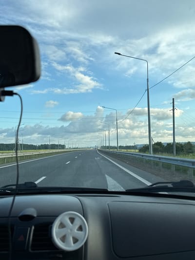 The image is taken from inside a vehicle, looking out through the windshield at a highway in Krasnokamsk, Russia. The road stretches into the distance, with multiple lanes marked by white lines. On the right side of the road, there is a metal guardrail, and beyond that, a grassy verge and trees. Streetlights line the highway on both sides, spaced at regular intervals. The sky is partly cloudy with fluffy white clouds against a blue background. The dashboard of the car is visible in the foreground, with a small, white, six-spoke decorative item attached to the air vent. A black rearview mirror is also visible in the upper left corner of the frame. The scene appears to be captured during daylight hours, with good visibility. There are no people or other vehicles immediately visible on the road. The overall impression is of a typical highway scene during a drive.