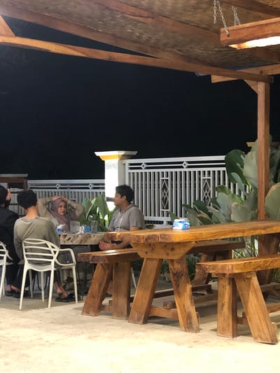 A group of people are gathered at an outdoor cafe at night. The setting appears to be rustic and casual, with wooden tables and chairs. Some of the people are seated at the tables, while others are standing or interacting. One person is leaning over a table, looking at something on it. Another person, wearing a hijab, is resting their head in their hands. The scene is dimly lit by artificial lights, creating a warm and inviting atmosphere. The background features a white fence and lush green foliage, suggesting a tropical location. The time of day is clearly night, with a dark sky visible. The overall mood seems relaxed and convivial. There is a bottle of water with text on it visible on a table.