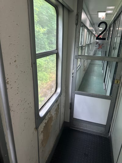 The image shows the interior of a train car. On the left side, there is a window with trees visible outside, suggesting a rural or forested area. The interior walls appear to be white with some discoloration and peeling paint. To the right of the window is a doorway with a glass panel. On the glass, the number "2" is prominently displayed in black. Below the "2" is a rectangular sign with a red circle and a diagonal line through a cigarette, indicating "no smoking." The corridor beyond the doorway appears long and narrow, with multiple doors on either side, suggesting passenger compartments. The floor is covered with dark, textured material, possibly rubber matting. The overall impression is of an older, functional train car. The location is Gornje Leskovice, Serbia.