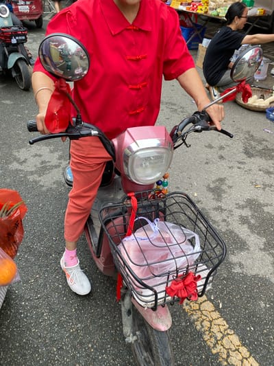 A person, seen from a high angle, is sitting on a red electric scooter in what appears to be a street or marketplace. The individual is wearing a vibrant red traditional Chinese jacket with frog fastenings and orange pants. White athletic shoes with pink socks are visible on their feet.

In front of the scooter, a black wire basket is attached, containing a clear plastic bag with pink items inside, possibly food. A red ribbon is tied to the basket, and a string of colorful beads hangs from it, adding a decorative touch. A round mirror on the scooter reflects another person, possibly the photographer, wearing glasses and a black t-shirt, looking away.

The scooter itself is a reddish-pink color with a silver handlebar and a large headlight. The ground is paved with asphalt and has yellow markings. To the left of the scooter, a metal cart or stall is visible, laden with goods in yellow and white packaging.

The scene suggests a daytime setting, likely in an urban environment given the pavement and marketplace elements. The overall mood is neutral, focusing on the everyday activity of riding a scooter, possibly for shopping or errands in Chongzhou City, China. There is no visible text in the frame.
