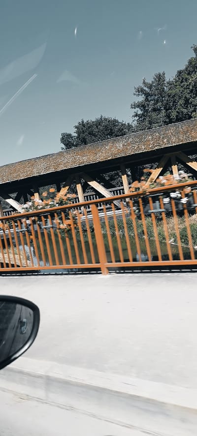 The image, captured from a moving vehicle, shows a long, covered wooden bridge with a rustic, weathered roof and visible wooden structural supports. An orange railing with vertical bars runs along the side of the bridge, adorned with flower boxes containing orange and white flowers. Below the railing, a body of water, likely a canal or small river, is visible with vegetation along its banks. In the foreground, the side-view mirror of the vehicle is partially visible on the left side of the frame, reflecting a distorted image of the surroundings. The sky above is a clear, light blue with some faint, elongated light streaks. The background features lush green trees. The image appears to be of the "Schulbergbrücke" (School Hill Bridge) in Winterbach, Germany, a well-known landmark often depicted with these aesthetic features.