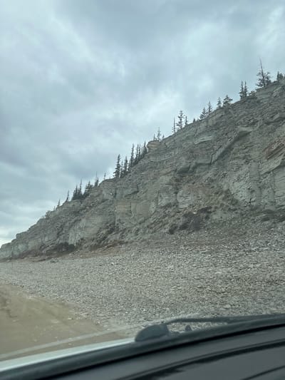 The image shows a cliff face beside a road, likely taken from inside a vehicle. The cliff is composed of gray rock with some vegetation (likely trees or shrubs) growing along its edge. Below the cliff, there is a sloping area covered in loose gravel and dirt, leading to the road. The sky is overcast with gray clouds. The vehicle window is visible on the right side of the frame. The exact location in the city of Bulgunnyakhtakh, Russia is unknown.