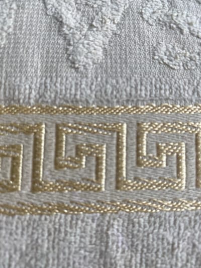 This media file is a close-up, high-angle shot of a textured textile, likely a towel or fabric with an embroidered pattern. The primary subject is the fabric itself, revealing its coarse texture with loops of thread. A prominent decorative element is a Greek key pattern, or meander, embroidered in a gold-colored thread. This pattern runs vertically along the right side of the frame, bordered by a braided gold thread. The background on the left of the frame shows a more abstract, raised texture, possibly a floral or organic design, also in a lighter, off-white shade. The lighting appears even, suggesting an indoor setting or overcast conditions, with no strong shadows indicating a specific time of day. There are no people or animals visible. The context provided indicates the location is Talas, Turkey, which might suggest this is a piece of traditional Turkish textile art or a modern item made in the region. The image does not contain any visible text. The overall impression is one of detail and texture, focusing on the craftsmanship and design of the fabric.