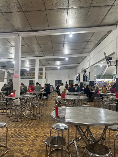 The image depicts an indoor eating establishment located in Nam Hải Lăng Commune, Vietnam. The scene is illuminated by multiple spherical LED lightbulbs mounted on a textured, grey tiled ceiling, which features exposed white beams. The floor is covered in brown and tan patterned tiles. White support pillars are spaced throughout the room, and several black wall-mounted oscillating fans are visible.
Approximately 25-30 individuals, primarily adults, are seated at numerous round and square stainless steel tables, utilizing matching backless stainless steel stools. Many tables feature red plastic caddies containing chopsticks and spoons, along with stacks of white ceramic bowls. Individuals are engaged in dining and social interaction; some are looking at their phones, while others converse.
In the mid-ground, a white pillar displays a partially obscured Vietnamese sign with the prominent text "ĐẶC SẢN" (Specialty), and further text indicating "RƯỢU MINH MANG, RƯỢU BÀU" (Minh Mang Wine, Bàu Wine), suggesting local specialties are offered. In the background on the right, a retail area or market stall is visible, displaying shelves with packaged goods and items hanging from racks. The overall environment suggests a casual, busy eatery or food court setting.