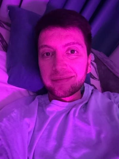 The media file is a close-up, horizontally oriented selfie of a person lying on a pillow in what appears to be a bedroom. The subject is a man with short, dark hair and stubble on his face. He is looking directly at the camera with a slight smile. His left eye is clearly visible, and his right eye is partially obscured. The image is illuminated with purple and pink lighting, creating a vibrant and somewhat surreal atmosphere.

The man is wearing a light-colored t-shirt, possibly light blue or gray, which is draped over his shoulder and chest. He is lying on a dark blue pillow, and behind him, there appears to be a patterned fabric, possibly bedding or a blanket. The background also shows a textured surface with vertical lines, which could be part of a wall or furniture, bathed in the same purple light.

The scene suggests a casual, relaxed moment, perhaps taken before sleep or upon waking. The lighting might indicate a mood setting or artificial light source. There is no visible text in the image. The location context is Tokaryovo, Russia, but this is not visually evident from the image itself. The overall impression is one of intimacy and personal reflection, captured in a low-light environment.