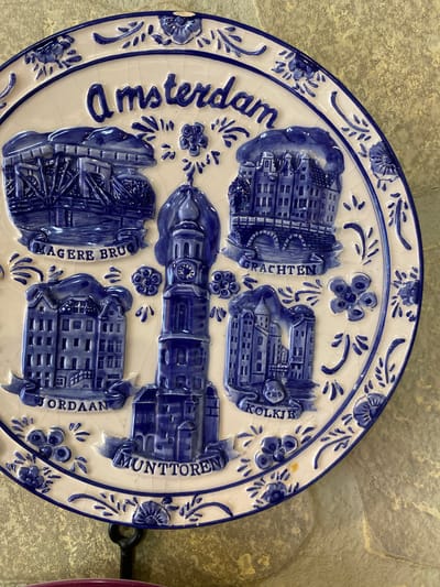 The image shows a close-up of a decorative blue and white ceramic plate featuring embossed depictions of Amsterdam landmarks. The plate is mounted on a wall with a textured, stone-like surface. The text "Amsterdam" is prominently displayed at the top in blue lettering. Below this, various buildings and architectural elements are depicted, each labeled with a blue banner. These include "Magere Brug," "Grachten," "Jordaan," "Munttoren," and "Kolkie." The plate is adorned with floral and decorative blue patterns around the rim. The background appears to be a neutral-colored, rough-textured wall. There are no people or animals visible in the image, nor any indications of the city of Yokohama, Japan.