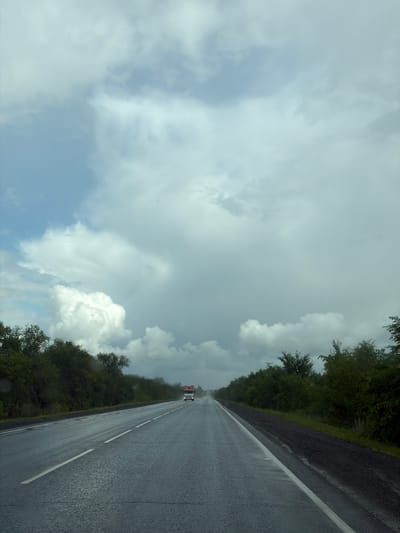The image depicts a two-lane asphalt road extending into the distance, flanked by dense green trees on both sides. The road surface is wet, indicating recent rain, with visible reflections and streaks of water. White lane markings are present, separating the traffic lanes. In the far distance, a white truck with red accents is visible, traveling in the same direction as the camera. The sky is predominantly filled with large, dramatic clouds, with patches of lighter blue sky showing through. The overall scene suggests a rural or semi-rural highway setting under overcast and potentially rainy weather conditions. There are no people visible in the frame.