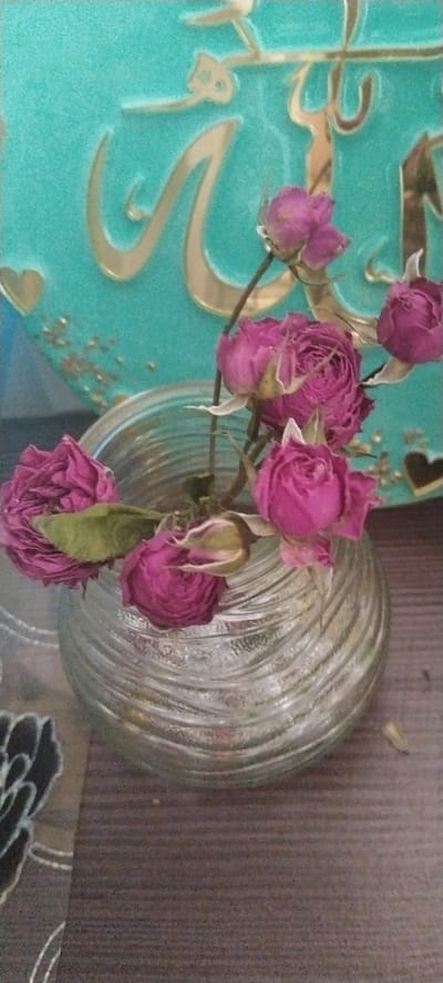 A still life composition features a clear glass vase with a swirling ribbed texture, filled with dried pink roses. The vase is placed on a dark wooden surface. In the background, a teal-colored decorative object with gold Arabic calligraphy and small gold heart shapes is visible. The overall lighting is dim, creating soft shadows. The image does not provide any information about the city of Aksu, Kazakhstan.