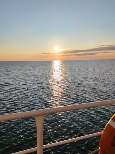 The image depicts a sunset over a body of water, viewed from the deck of a vessel. The sun is low on the horizon, casting a golden reflection across the rippling surface of the sea. Wispy clouds are visible in the pale sky, tinged with orange and yellow from the setting sun. In the foreground, a white railing runs horizontally across the frame, with a portion of an orange life preserver visible on the right side. The water appears dark and deep, with small waves creating a textured surface. The scene is serene and suggests a calm atmosphere. There are no people or specific city landmarks visible to indicate it is in Kakkarovo, Russia.