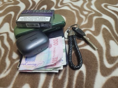A close-up, overhead shot on a brown and white striped blanket shows several items: a dark grey "hoco." branded earbud case, a pack of "TEREA" brand cigarettes for IQOS ILUMA, a green tobacco heating device, a dark blue wallet or card holder, several Ukrainian Hryvnia banknotes, and a set of keys attached to a black braided leather fob. The banknotes include a prominent 100 Hryvnia note, and another partially visible note with the denomination "200". The cigarettes pack displays warning text in Ukrainian. The scene appears to be indoors, on a blanket, with no identifiable urban features or references to the city of Khotsky, Ukraine.
