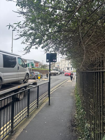 This image depicts an overcast daytime street scene at St Paul's Roundabout in Salisbury, United Kingdom.
In the foreground, a black metal pedestrian barrier lines the left edge of an asphalt sidewalk. To its right, a black pole supports a square black sign with white graphics. Overhanging branches with green leaves partially obscure the upper left corner of the frame. On the far right, a tall, dark metal fence, partially overgrown with ivy, borders the sidewalk.
The midground features a two-lane asphalt road with double yellow lines painted along the curb. On the left lane, a silver van, appearing to be a Renault Trafic, is loaded onto a flatbed recovery truck. The van's front wheels are secured with a chain. Behind the recovery truck, a dark grey SUV with hazard lights illuminated is positioned near a yellow 'keep left' bollard. Further down the road, a queue of vehicles includes a white flatbed truck carrying material. In the right lane, a red hatchback is approaching the camera. A single individual walks away from the camera on the right-hand sidewalk, wearing dark clothing and carrying a bag.
The background reveals a large green directional sign for "St Paul's Roundabout," indicating routes to "Romsey A3090," "Stockbridge A30," and "Southampton A36." Beyond the road, white, multi-story terraced buildings are visible under an overcast sky.