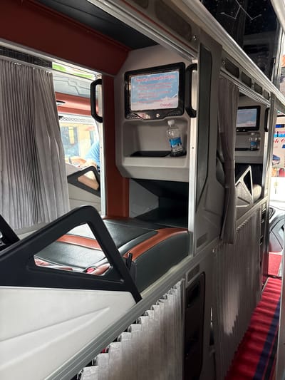 The image shows the interior of a bus or sleeper train, likely in Lao Bảo Commune, Vietnam. The scene is focused on the passenger seating and sleeping compartments. In the foreground, a black and orange upholstered seat with a black armrest is visible. To the left, a grey pleated curtain is drawn. Behind the seat, a small monitor displays text in what appears to be Russian, with a snowflake graphic and the word "Спасибо" (Thank you). Below the monitor is a shelf holding a bottle of water. Further back, another similar compartment is visible with a drawn grey curtain, another small monitor, and a shelf with a bottle of water. The overall impression is one of compact, private sleeping or resting areas within a vehicle. The flooring is a red carpet with dark blue and red stripes. The environment appears to be indoors.