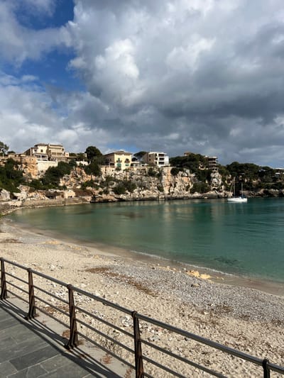 The image depicts a coastal inlet in Porto Cristo, Spain, under a partially cloudy sky featuring dark grey storm clouds transitioning to lighter white clouds and blue sky. The foreground comprises a grey flagstone walkway bordered by a dark, weathered metal railing with horizontal bars, casting shadows to the left. Beyond the railing, a sandy beach mixed with small light-colored pebbles extends to the calm, turquoise-green water of the inlet. On the far side of the inlet, light-colored rocky cliffs rise, topped with green vegetation and numerous multi-story buildings, predominantly beige, white, and light yellow, exhibiting varied architectural styles. A single white sailboat with a mast is anchored in the water towards the right side of the inlet, near the cliff face. No individuals are visible.