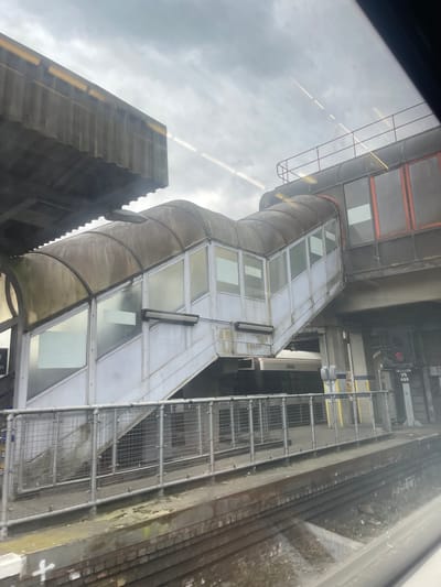 The image depicts a view of Sevenoaks railway station in the United Kingdom. Visible in the shot is the exterior of an elevated passenger walkway connecting different platforms. The walkway is constructed of metal and glass panels. Adjacent to the walkway, there is a metal ladder-like structure running alongside the brick exterior of the rail infrastructure. In the background, there are buildings of the city, and overhead, the sky is overcast.