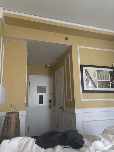 The image depicts an interior scene of a hotel room. The walls are painted a soft yellow with white decorative molding. On the left side of the image, a white lamp is partially visible against the yellow wall. Further to the left, a brown paper shopping bag with green text is placed on the floor. The bag appears to contain items and has handles. In the center of the image is a closed white door, likely the room's entrance, with a metal plate and a key card reader. A door handle and a vertical metal fixture are visible on the door. To the right of this door, another white door is partially visible. Above the doors, the ceiling is white. On the right wall, a framed black and white picture hangs, featuring multiple panels with images of architectural elements, possibly the Eiffel Tower, and natural scenery. Below the picture frame, the wall transitions to white wainscoting. A dark-colored garment is draped over a white surface in the foreground, which appears to be a bed. The lighting suggests an indoor environment. The image is taken from a low angle, looking upwards. The scene is static and shows no people. There is no discernible information in the image to suggest the specific location within San Francisco, United States of America.