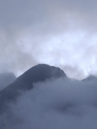 The image depicts a mountain peak shrouded in mist and clouds. The sky is overcast with a uniform grey tone, suggesting a cloudy or foggy day. The mountain itself is dark and silhouetted against the lighter sky. Wisps of fog or low-lying clouds obscure the lower slopes of the mountain and surround it, creating a sense of depth and atmosphere. The scene is devoid of any human presence or artificial structures. The environment is natural and rugged, characteristic of a mountainous region.