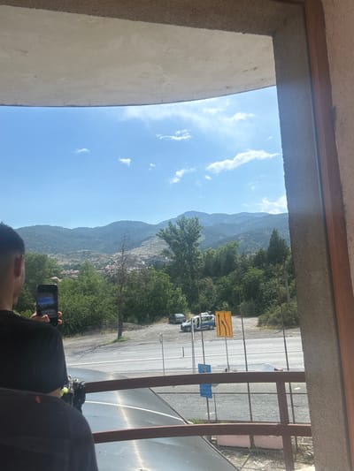 The image captures a scenic view of a mountainous landscape, likely in or near Simitli, Bulgaria, as suggested by the provided information. The foreground is partially obscured by a textured concrete overhang and a wooden frame, suggesting the vantage point is from a building's interior or balcony.
In the mid-ground, a person is visible from the back, holding a smartphone and appearing to take a photograph or video of the surrounding scenery. The person is wearing a black t-shirt. Beyond this individual, a road runs horizontally across the frame, with a white police vehicle and a yellow traffic sign with red arrows indicating lane closures. Dense green trees and vegetation cover the lower slopes of the hills, with some buildings and houses visible nestled within the landscape. The background is dominated by rolling mountains under a clear blue sky with scattered white clouds. The overall impression is one of a rural or semi-rural setting with natural beauty.