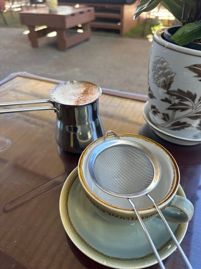 The image depicts a close-up view of a coffee setup on a dark, reflective table, likely outdoors. In the foreground, a light blue teacup and saucer are positioned, with a silver mesh strainer resting on top of the cup. To the left, a small, metallic coffee pot, known as a cezve or ibrik, contains a frothy coffee beverage, dusted with cinnamon or cocoa powder. In the background, out of focus, a wooden table and chairs are visible, suggesting a cafe or outdoor seating area. A potted plant in a decorative ceramic pot is situated to the right of the coffee pot. The lighting suggests it is daytime, with sunlight filtering through. The scene is likely set in Bellingen, Australia, given the context.