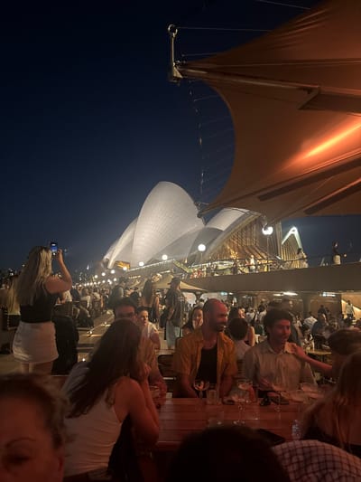 A group of people are seated at a long wooden table, likely dining at a restaurant. Sydney Opera House is visible in the background. The scene is illuminated by artificial light, with an orange sail-like structure above the dining area. A person is taking a photo of the Opera House with their phone.