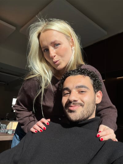 The scene depicts two individuals, one female and one male, in an indoor setting located in what is indicated as Marche-lez-Écaussinnes, Belgium. No specific identifying landmarks within the city are visible in the indoor environment.

The female, positioned behind the male, has long blonde hair, light skin, and blue eyes. She is wearing a dark brown long-sleeved sweater and blue jeans. Her hands, with fingernails painted red, rest on the male's shoulders. She has pursed lips and looks directly at the camera.

The male, in the foreground, has dark curly hair, a beard, and a mustache, with light-to-medium skin and brown eyes. He is wearing a black turtleneck sweater and smiles towards the camera.

Both individuals are illuminated by strong natural light originating from the left side of the frame, casting shadows. The background includes light-colored rectangular panels affixed to the ceiling and indistinct darker architectural features. On the far left, a blurred surface holds objects consistent with a camera or electronic device and glassware.