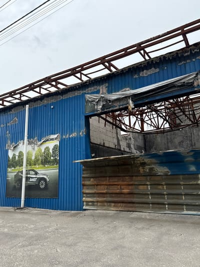 The image shows a partially destroyed building in Horenka, Ukraine (50.56, 30.309). The building is made of blue corrugated metal and has a rusty metal roof. The roof has been partially destroyed, and the building appears to have been damaged by fire or an explosion. The image shows a black car poster on the building, the poster features a black car in the foreground, and trees in the background, with the word "BANDIT" on the side of the car. The building is located in a residential area, and there are no people present. The building is surrounded by rubble and debris. The image shows the destruction caused by the Russian invasion of Ukraine.