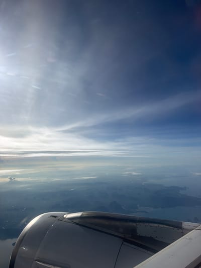 The image is an aerial view from an airplane, showing a landscape below and the sky above. In the foreground, the engine nacelle of the aircraft is visible, taking up the lower left portion of the frame. The nacelle is metallic and curved, with visible seams and rivets. Beyond the aircraft, a vast expanse of sky stretches across the upper half of the image. Wispy clouds are present, with a bright light source, likely the sun, illuminating the scene from the left, creating lens flares and streaks of light.
Below the clouds, a hazy landscape unfolds. It appears to be a coastal region with numerous islands or mountainous terrain emerging from the water. The landmasses are depicted in shades of blue and grey, suggesting a significant distance and possibly atmospheric perspective. Rivers or inlets of water are visible winding through the terrain. In the far distance, on the left side of the landscape, a plume of smoke or ash is rising from what appears to be a volcano.
The image does not depict any people, specific actions, or interactions. It is a static, environmental shot from a high altitude. The scene is not identifiable as being specifically in Sisa Chorakhe Noi Subdistrict, Thailand, as there are no landmarks or distinguishing features to confirm the location. The general appearance is consistent with a tropical or subtropical coastal area with volcanic activity.