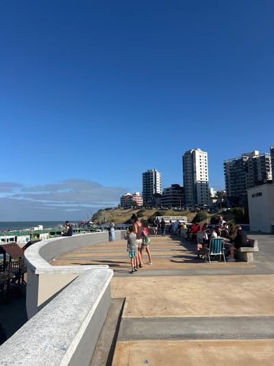 A sunny day at a coastal city. Several tall buildings line the coastline. People are walking, sitting, and enjoying the view from a concrete promenade. A low wall runs along the edge of the promenade, separating it from the beach. The sky is clear and blue. The sea is visible in the distance.