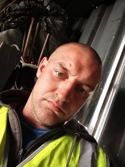 This is a close-up, high-angle, selfie-style photo of a man with a shaved head and stubble. He is wearing a bright yellow and gray high-visibility jacket over a blue t-shirt. The jacket is partially unzipped, revealing the t-shirt underneath. The man has a serious expression on his face, with his eyebrows slightly furrowed and his lips in a slight frown. His eyes are open and looking towards the camera. There is a small bump on his forehead.
The background is dark and appears to be an industrial or storage setting. To the left, there are black plastic bags and some metallic objects that look like rebar. To the right, there is a dark, corrugated surface, possibly part of a container or vehicle. The lighting is somewhat harsh, creating strong shadows on the man's face. The overall impression is one of a candid, perhaps work-related, moment captured in a somewhat rugged environment. There is no visible text in the image. The location context provided is Raygorodok, Kazakhstan.