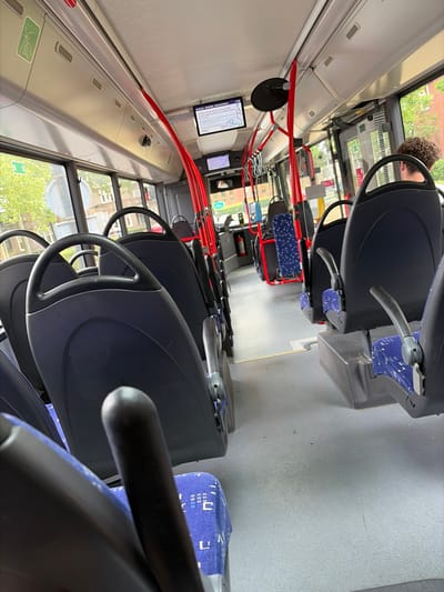 This media file depicts the interior of a bus in Enschede, Netherlands. The camera is positioned looking forward and slightly upwards, capturing a row of empty dark blue bus seats with blue patterned fabric inserts. The seats have grey plastic casings and silver handles. Red poles, likely for passenger support, run along the length of the bus, visible in the foreground and midground. A black circular mirror is mounted on the left side of the bus.
A small screen displaying a map or route information is visible on the left wall of the bus. The windows along the right side of the bus show a blurry view of trees and buildings, suggesting an urban or suburban setting. The lighting appears to be natural, indicating daylight, likely mid-morning or afternoon. The overall scene is one of an empty public transport vehicle, possibly between stops or at the beginning/end of a route. No people are clearly visible, though a portion of a person's head and shoulder, with curly brown hair, is present at the top left of the frame, suggesting someone might be the bus driver or a passenger in the front. There is no discernible activity taking place other than the implied movement of the bus. The weather outside appears to be clear with green foliage visible. A green sign with white text in a stylized font is visible on the left wall.
