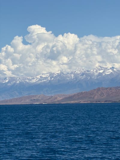 The image shows a landscape oriented vertically, featuring a bright blue sky with large white cumulus clouds on the left. Next to the clouds are snow-capped mountains that transition into brown mountains on the right. A narrow strip of land is visible at the base of the brown mountains. To the right of the land is the dark blue water of Lake Issyk-Kul in the city of Tong, Kyrgyzstan. Ripples on the water's surface suggest a slight breeze. No people are visible.