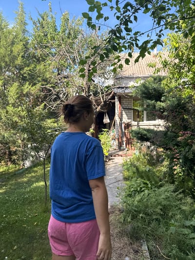 A woman with her back to the camera, wearing a blue t-shirt and pink shorts, stands on a pathway in a yard. She has her hair pulled up in a bun. The yard is overgrown with various plants and trees, and a small house with a white brick facade and a grey, corrugated roof is visible in the background. The scene appears to be in Balivka, Ukraine, judging by the surrounding vegetation and architectural style of the house, consistent with a residential area in the region. The pathway is made of concrete or stone slabs. The overall impression is a somewhat unkempt but lived-in garden setting.