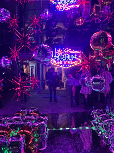 The image appears to be a photograph taken in a mirrored environment, possibly an entertainment venue, featuring various decorative elements. Prominent features include multiple metallic balloons (spherical and star-shaped), and neon signs with the words "Maison Welcome to Fabulous Las Vegas Nevada". The mirror reflections create a kaleidoscopic effect, with reflections of people visible in the background. The scene is illuminated with purple and pink hues, adding to the vibrant atmosphere. No location in Ezeiza, Argentina is discernible in the photograph.
