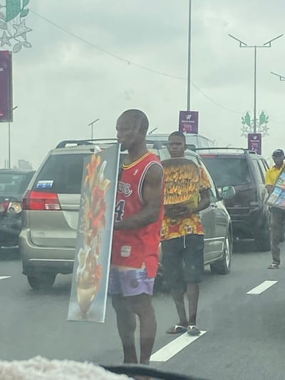 The image depicts an urban street scene in Ebute-Metta, Nigeria, observed from inside a vehicle. The setting is a paved road under an overcast sky, with multiple vehicles stationary or moving slowly in traffic.
In the foreground, slightly to the left, stands a dark-skinned adult male with a shaven head, facing left in profile. He wears a red sleeveless basketball jersey with "76ers" and the number "4" visible, paired with purple patterned shorts. He holds a large, rectangular framed artwork vertically against his left side, showcasing an abstract or floral design in orange, red, and gold tones.
Behind him and slightly to the right, another dark-skinned adult male is visible from the waist up, facing forward. He wears a yellow and dark-patterned short-sleeved shirt and dark shorts, with white flip-flops visible on his feet. He holds a large, flat, circular item, densely covered with what appears to be dried fish or similar food items.
Further in the background, to the far right, a third dark-skinned adult male is partially visible, wearing a yellow shirt and a dark baseball cap, carrying multiple packaged items.
The background shows several parked or slow-moving cars, including a light-colored SUV directly behind the first two individuals. White solid and dashed lane markings are visible on the asphalt road surface. Overhead, streetlights and decorative elements, including green star and foliage motifs and purple banners with partially obscured text, are attached to poles, suggesting public infrastructure and possibly a festive period. The individuals appear to be street vendors engaged in hawking goods to occupants of vehicles.