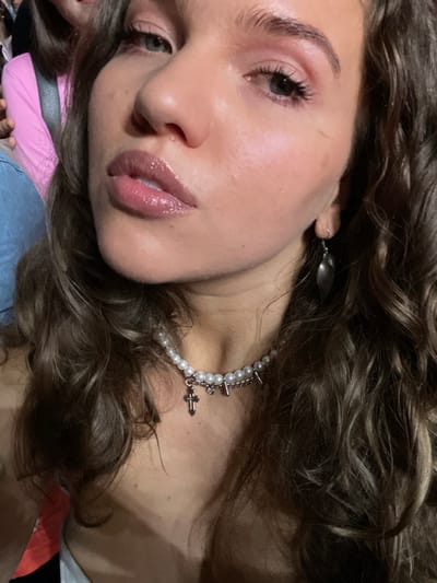 A woman with curly brown hair is seen up close, gazing into the camera. She has fair skin, full lips with glossy lipstick, and delicate makeup highlighting her eyes. She wears a pearl necklace with spikes and a cross pendant, plus a simple silver earring. The photo is taken at a low angle.
