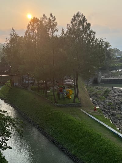 This image depicts a serene outdoor scene at a resort or park, possibly located near Krasak, Indonesia, as suggested by the presence of lush greenery and tropical trees. The sun is setting, casting a warm glow over the landscape, with the sun partially obscured by tall pine trees in the foreground.
The scene features a children's playground with a yellow slide and colorful climbing structures, set on a grassy area. Nearby, there are several umbrella-covered tables, suggesting a dining or relaxation area. A wooden walkway or bridge extends from the left side of the image, leading towards the playground and seating area.
A narrow, calm river or canal runs along the left side of the image, with a stone embankment. On the right side of the image, a rocky riverbed leads to a concrete dam or spillway, with water flowing over it. A person can be seen fishing in the river near the dam. The overall environment is natural and tranquil, with a mix of cultivated greenery and rugged, natural elements.