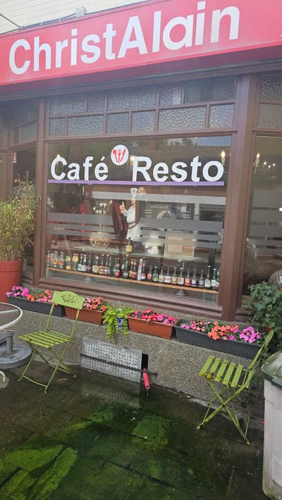 This image captures a street-level view of the exterior of a cafe or restaurant named "ChristAlain Café Resto" in Thuin, Belgium. The scene appears to be set during the daytime, with natural light illuminating the facade.
The building features a prominent red awning at the top with "ChristAlain" written in large white capital letters. Below the awning, there's a large display window with brown frames, part of which has textured glass panels. Across the main window, "Café Resto" is displayed in white text on a purple horizontal stripe, accompanied by a small icon of a fork and knife.
Through the window, various bottles are visible, neatly arranged on shelves inside the establishment. A cutout or poster of a chef figure, dressed in white, is also prominently displayed inside, giving a thumbs-up gesture.
Outside the cafe, there's a small seating area. Two light-green metal folding chairs are placed on the pavement. Several flower boxes filled with vibrant pink and orange flowers add a splash of color. A large red planter with bamboo-like plants is visible on the far left. The ground is paved but appears damp, with noticeable patches of green moss or algae, especially in the foreground and towards the right, suggesting a moist environment or recent rain. A metal grate and a red pipe are also present on the ground near one of the chairs. No direct human activity is depicted in the static scene.