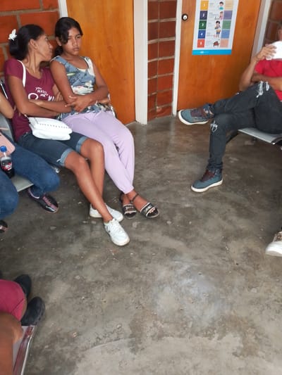 The scene depicts an indoor waiting area, possibly in a clinic or public building. Two women sit closely together on a metal bench, the woman on the left wearing a maroon t-shirt, denim shorts, and white sneakers, while the woman on the right wears a sleeveless print top and light purple pants. To the right of the women, a man sits with his legs crossed, wearing a red shirt and ripped black jeans with blue sneakers. Another person is partially visible to the left of the two women.

A handwashing instruction poster is visible on the orange wall behind the seated man. The floor is a gray-toned concrete. There are no clear visual cues to definitively identify the location as Barcelona, Venezuela.