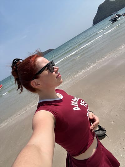 A woman with auburn hair in a bun wears sunglasses and a burgundy t-shirt with white lettering on a sunny beach. The tide laps at the sand. A knapsack sits on the sand near her. In the background, people gather near boats at the water's edge, and a landmass juts into the water. The sky is clear and blue.
