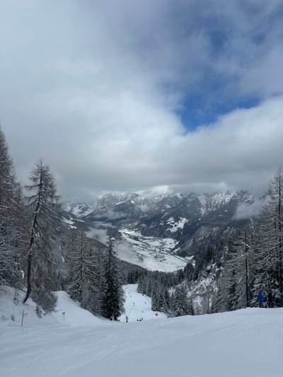 The image captures a panoramic winter view from a ski slope in Bad Hofgastein, Austria. The foreground displays a wide, groomed ski trail covered in extensive snow, exhibiting visible ski tracks. Multiple skiers are present on the lower section of the slope, engaged in descent or standing; one skier in a blue jacket is partially visible on the right edge. Orange marker poles are positioned along the left boundary of the trail. The slope is bordered by dense forests of snow-laden pine and larch trees. In the mid-distance, a snow-covered valley is visible, containing a town with discernible buildings and a winding road or waterway, partially obscured by low-lying mist. The background features a range of rugged, snow-capped mountains, intermittently veiled by heavy clouds. The sky above presents a mix of overcast grey clouds and distinct patches of clear blue.