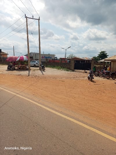 The image shows a dirt road with a paved road on the left. There is a power line running across the top of the image. There are multiple motorcycles parked along the side of the dirt road, and a white car parked near the power line. There are several people near the motorcycles, and two more people are sitting at a table under a pink and white umbrella. A building with the word "drilander" painted on its side is visible in the background. The location of the image is in Okinni, Nigeria, at the coordinates 7.832 latitude and 4.51 longitude. The weather is sunny with a few clouds in the sky.
