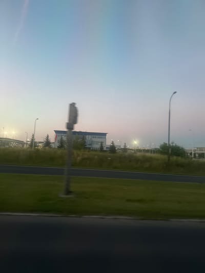 The image depicts a street scene at dusk in Lublin, Poland. In the foreground, a dark asphalt road is visible, bordered by a strip of green grass. A concrete bollard stands in the middle of the grassy area. Behind the grass, a row of trees, some appearing to be evergreen, are sparsely planted. A modern, multi-story building with a flat roof and numerous windows is situated in the middle ground. A Polish flag, red and white, is affixed to the side of this building. Numerous streetlights are present throughout the scene, some illuminated, casting a soft glow. In the background, more buildings and structures are visible, along with a bright, large streetlight. The sky is a gradient of light blue and pale pink, indicating the twilight hours. The overall impression is one of an urban or suburban periphery, possibly an industrial or commercial zone, as suggested by the modern architecture and infrastructure. There are no people visible in the frame.