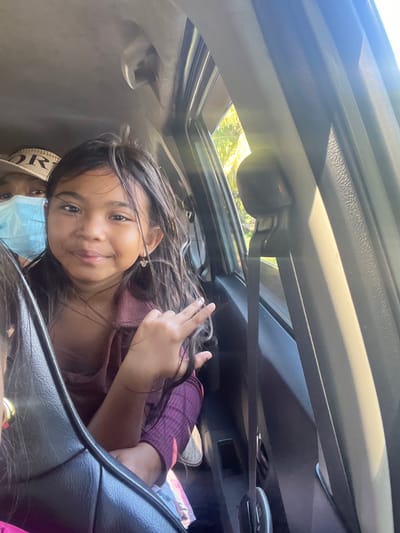 A young girl with dark hair and a light complexion smiles and makes a peace sign with her right hand while riding in a car. She wears a pink top and a small gold earring. Another person, wearing a light blue mask and a hat with the word "Dior" on it, is partially visible behind her. The interior of the car is visible, including a black seatbelt and a dark gray door panel. The image appears to be taken during the daytime, with sunlight filtering through the car window, casting shadows and highlighting the interior. The environment outside the car suggests a warm, sunny day, with glimpses of green foliage and bright sunlight. The context places this scene in the city of Gerokgak, Indonesia.
