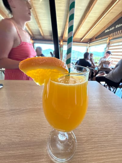 A close-up shot focuses on a glass of orange juice garnished with an orange slice, placed on a wooden table. Two green and white striped straws extend from the glass. In the background, through a window, several people are visible sitting at tables in what appears to be a restaurant or cafe. The setting features wooden beams and a ceiling with slatted panels, suggestive of an indoor, possibly rustic, establishment. Sunlight streams in, indicating daytime. The overall mood is relaxed and casual. A portion of a person wearing a pink and white striped top is visible in the lower left of the frame. Visible text on a dark banner in the upper background reads "Dobro došli - Welcome". The location is identified as Miločani, Montenegro.