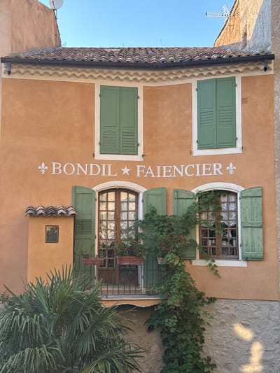A two-story building with an orange facade and a tiled roof is depicted in Moustiers-Sainte-Marie, France. White lettering on the facade reads "BONDIL FAIENCIER," with a fleur-de-lis symbol on either side. Two green-shuttered windows are visible on the upper floor. On the lower floor, there is a balcony with a black metal railing, flanked by two green-shuttered windows. The window on the left has an open shutter and a small, square opening above it covered with roof tiles. A small painting is visible within this opening. A palm tree is in the foreground on the left, and green vines with red flowers are growing up the facade around the right lower window. The lower part of the building is a stone wall. The sky is clear and blue.